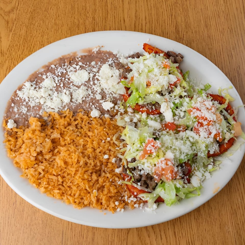 Mexican platter with red rice, refried beans, grilled meat, lettuce, tomatoes, and crumbled cheese on a white plate.