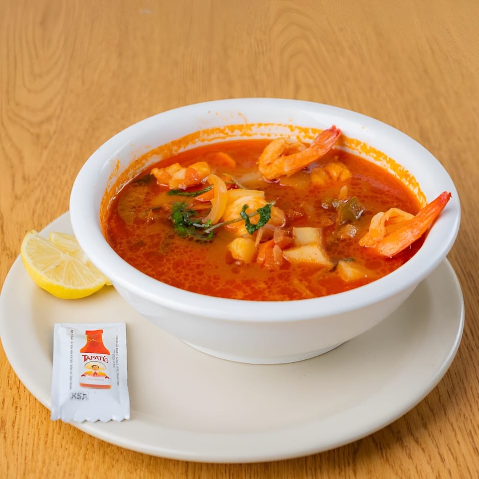 Red shrimp soup with vegetables, a lemon wedge, and Tapatio hot sauce packet on a wooden table.